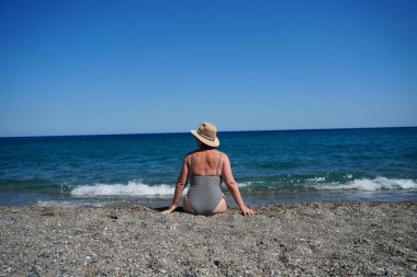 Kadın okyanusa bakan çakıl taşlı bir plajda oturuyor. Şapkası ve desenli mayosu var. Arka planda dalgalar ve yukarıda açık mavi gökyüzü ile rahatlatıcı an