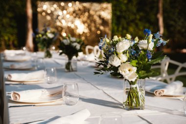 Açık hava yemek masası beyaz çarşaflar ve cam süslemelerle süslenmiş, taze çiçeklerle süslenmiş. Arka planda bulanık altın ışıklar sakin bir atmosfer yaratır..