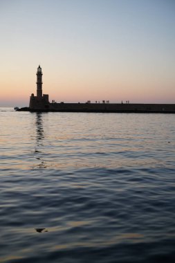 Gün batımında bir deniz fenerinin silueti. Sakin su ve ufukta uzak figürler. Gökyüzü sıcaktan serin tonlara geçerek dingin ve sakin bir atmosfer yaratıyor..