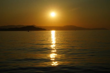 Güneşin sakin bir denizin üzerinde battığı sakin günbatımı sahnesi suya altın yansıyor. Uzak dağların siluetleri huzurlu ve huzurlu bir ortam yaratır.