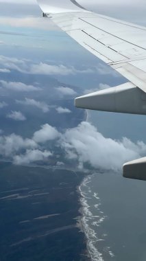 Aşağıdaki sahil şeridini ve bulut oluşumlarını gösteren bir uçak penceresinden manzara. Uçakların kanatları belirgin bir şekilde görünür, uçuş ve seyahatin özünü yakalar..