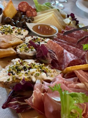 Gurme şarküteri kurulunda çeşitli peynirler, pişirilmiş etler ve zeytinler, ahşap bir tabakta sanatsal olarak sıralanmış. Fıstık ezmeli ekmek dilimleri gösteriyi güzelleştirir..