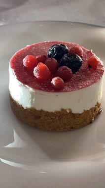 Kremalı bir cheesecake, üzerinde canlı böğürtlenler olan beyaz bir tabağın üzerinde zarif bir şekilde oturur. Tatlının zengin dokusu ve renkli meyve garnitürü iştah açıcı bir sunum oluşturur..