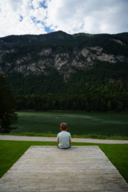 Genç çocuk sakin bir dağ gölünün yanındaki ahşap platformda sessizce oturuyor, yemyeşil ve yüksek zirvelerle çevrili, doğanın güzelliğini ve sükunetini yansıtıyor.