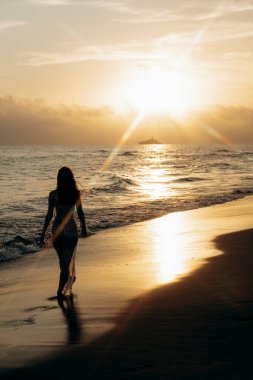 Gün batımında sakin bir sahil boyunca yürüyen bir kadın, canlı gökyüzüne karşı siluet çiziyor. Sahne, okyanusun huzurunu ve batan güneşin güzelliğini yakalar.