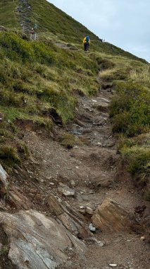 Maceracılar yeşil bir dağ yamacında dik ve kayalık bir yolda ilerlerler. Grup, açık hava keşiflerinin zorluklarını ve güzelliklerini vurgulayarak azimle yükseliyor..