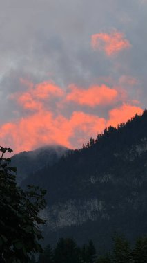 Gün batımında dingin bir dağ manzarasında canlı turuncu bulutların büyüleyici görüntüsü. Ormandaki dağın silueti renkli gökyüzü ile çelişiyor ve çarpıcı bir doğal manzara yaratıyor..