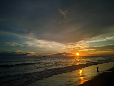 Huzurlu bir sahil sahnesi. Gün batımı sahnesi. Sahil boyunca yürüyen bir çocuk, dingin doğal güzelliği yansıtıyor.
