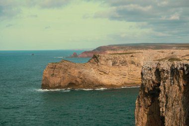 Bulutlu bir gökyüzünün altında mavi okyanusun üzerinde yükselen engebeli kıyı uçurumlarının manzarası.