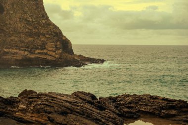 Bulutlu bir gökyüzünün altında mavi okyanusun üzerinde yükselen engebeli kıyı uçurumlarının manzarası.