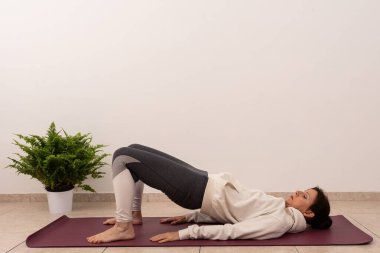 Bir kadın mindere yoga pozu veriyor. Siyah pantolon ve beyaz bir kapüşonlu giyiyor.