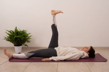 Bir kadın bacak bacak üstüne atmış bir şekilde paspasın üzerinde yatıyor. Beyaz kapüşonlu ve gri pantolonlu.