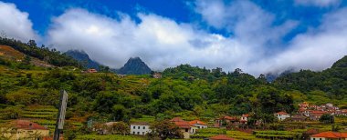 Parlak evler ve yamaçlarda sebze bahçeleri olan bir dağ köyü. Tropik bir adada açık havada bulutlarda gizlenmiş dağ zirveleri.   