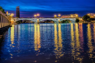 Triana Köprüsü gece karanlığında Sevilla, Endülüs, İspanya 'daki Guadalquivir Nehri' nin yansımalarıyla aydınlandı..