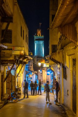 Bir grup insan, Fas 'taki Fez Medinesi' nde, Medersa Bou Inania minaresi yakınlarındaki dar bir ara sokakta gece ayakta duruyor..