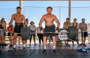 Sporcular Seville, İspanya 'da düzenlenen yoğun bir CrossFit yarışmasında halter gösterisi yapıyorlar. Kalabalık katılımcıları izliyor ve alkışlıyor.