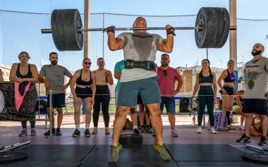 Sporcular Seville, İspanya 'da düzenlenen yoğun bir CrossFit yarışmasında halter gösterisi yapıyorlar. Kalabalık katılımcıları izliyor ve alkışlıyor.