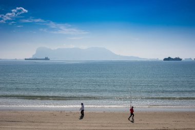 İspanya 'nın Algeciras kentindeki El Rinconcillo plajında yürüyen ve koşan insanlar, uzak arka planda kargo gemileri ve Cebelitarık ile.