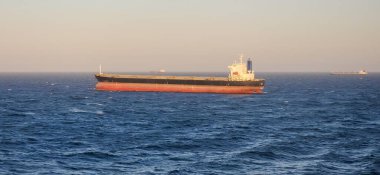 Büyük bir kargo gemisi, siyah bir gövdesi ve sakin mavi bir denizde yüzen kırmızı bir kıçı var. Uzak gemiler gün batımında açık, pastel renkli gökyüzünün altında görülebilir..