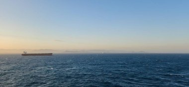 Büyük bir kargo gemisi berrak bir gökyüzünün altında sakin mavi bir denizde yelken açar. Uzak dağlar ve ufukta başka bir gemi görünüyor. Yumuşak güneş ışığı manzarayı aydınlatıyor..
