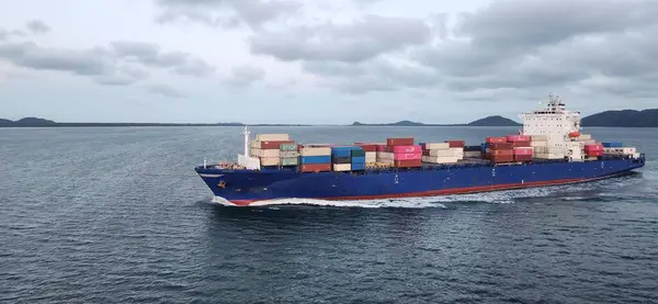 Stock image A large blue cargo ship carrying multicolored shipping containers sails through calm waters. Cloudy sky and distant islands in the background.