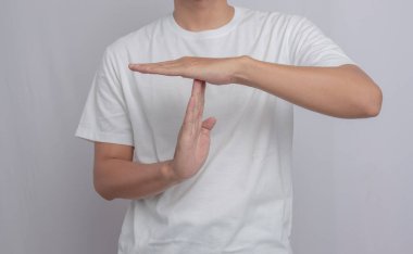 Sıradan giyinmiş sakallı genç bir adam beyaz bir arka planda 'mola' ya da 'duraklama' hareketi yapıyor.