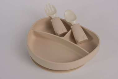 Açık kahverengi silikon bebek plakası, beyaz arka planda izole bir kaşık ve çatalla. Bir bebeğin ilk beslenmesi konsepti için bir araç.