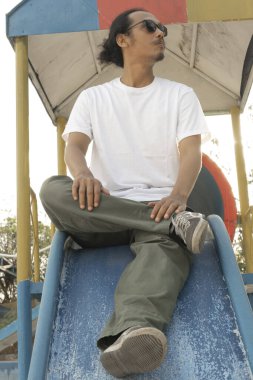 Sade beyaz tişört ve siyah güneş gözlüğü giyen Asyalı bir erkek model, gündüz vakti ticari bir parkta poz veriyor. Erkek mankenin beyaz tişörtü vitrin için model nesne olarak kullanılıyor.