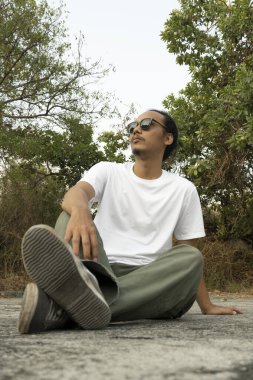 Sade beyaz tişört ve siyah güneş gözlüğü giyen Asyalı bir erkek model, gündüz vakti ticari bir parkta poz veriyor. Erkek mankenin beyaz tişörtü vitrin için model nesne olarak kullanılıyor.