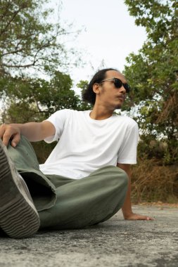 Sade beyaz tişört ve siyah güneş gözlüğü giyen Asyalı bir erkek model, gündüz vakti ticari bir parkta poz veriyor. Erkek mankenin beyaz tişörtü vitrin için model nesne olarak kullanılıyor.