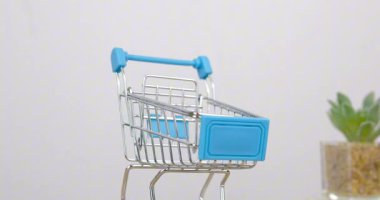 Beyaz masa arkasındaki metal bir alışveriş arabası yavaşça dönüyor. Dönen masada mavi ve krom renkli alışveriş sepeti.