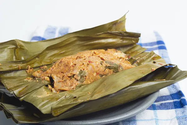 Pepes Tahu Jamur, baharatlı tofu ve mantar, muz yapraklarına sarılı, sonra buğulama ve ızgara. Ahşap masa veya motif tabakta servis ediliyor.