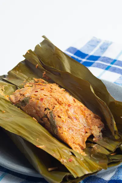 Pepes Tahu Jamur, baharatlı tofu ve mantar, muz yapraklarına sarılı, sonra buğulama ve ızgara. Ahşap masa veya motif tabakta servis ediliyor.