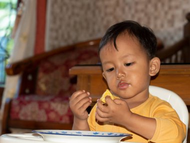 Toddler 'ın tost yerken yakın çekim. Mutlu Asyalı çocuk sandalyesinde peynir ve yumurta ile tost kahvaltısı yapıyor. Küçük çocuk kahvaltısını yüksek sandalyesinde ve masasında yapıyor..