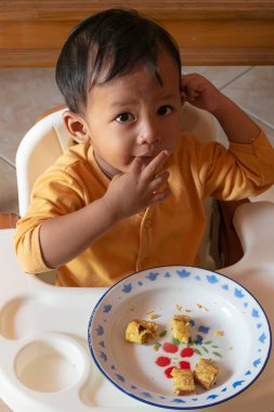 Toddler 'ın tost yerken yakın çekim. Mutlu Asyalı çocuk sandalyesinde peynir ve yumurta ile tost kahvaltısı yapıyor. Küçük çocuk kahvaltısını yüksek sandalyesinde ve masasında yapıyor..