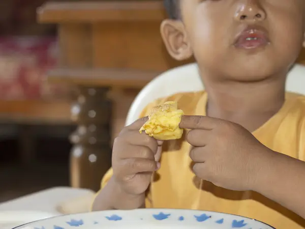 Toddler 'ın tost yerken yakın çekim. Mutlu Asyalı çocuk sandalyesinde peynir ve yumurta ile tost kahvaltısı yapıyor. Küçük çocuk kahvaltısını yüksek sandalyesinde ve masasında yapıyor..