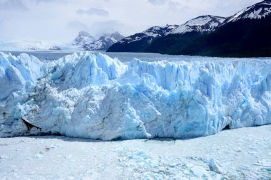 Patagonya 'daki Perito Moreno buzulunun önünde kristal mavi buz oluşumları ve arka planda karla kaplı kayalık dağlar var.