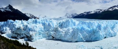 Patagonya 'daki Perito Moreno Buzul Cephesi' nin geniş ekran görüntüsü. Arka planda kristal mavi buz oluşumları ve karla kaplı kayalık dağlar var.