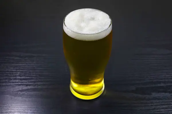 İçi bira dolu, köpüklü bir katman köpükle kaplı, parlak siyah bir masanın üzerine kurulmuş bir bardağın yakın çekimi. Görüntü, biranın zengin dokusuna ve kabarcıklarına odaklanır ve koyu renkli masa camı vurgulayan çarpıcı bir kontrastı sağlar. Öge