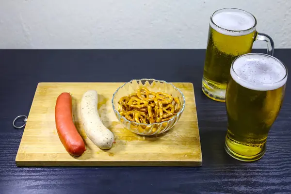 İki geleneksel Alman sosisi, kristal bir kasede gevrek krakerler, dolu bir bira bardağı ve bir bira bardağı, hepsi de ahşap bir tepside. Alman mutfağı ve Ekim Festivali kutlamaları için ideal..