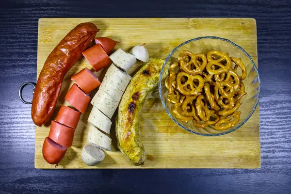 Yukarıdan yakından bakınca, iki Alman sosisi, iki dilimlenmiş ve iki bütün, kristal bir kasede altın çıtır krakerler eşliğinde, hepsi de kırsal bir ahşap tepside sunuluyor. Bu kurulum klasik ve davetkar bir Alman yemeğini ön plana çıkarıyor.