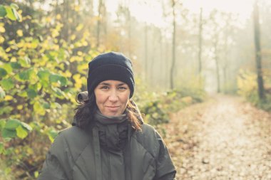 40-50 yaşlarında, ormanda gülümseyen bir kadın, izleyiciye bakıyor, soğuk Kasım sonlarında sıcak bir şekilde giyinmiş. Dijital detoks kopukluğu, sağlıklı yaşam tarzı, soğukkanlılıkla yürür. Samimi. Boşluğu kopyala