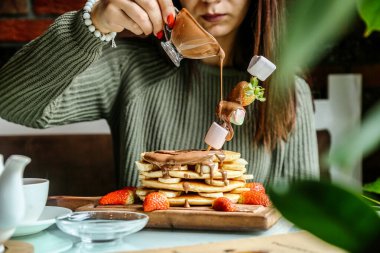 Bir kadın beyaz bir tabakta uzun bir yığın altın kahverengi krep üzerine şurup döker..