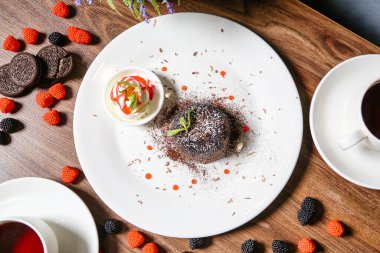 Üzerine lezzetli bir tatlı konmuş beyaz bir tabak dumanı tüten bir fincan kahvenin yanında oturur..