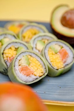 Mavi tabağın üzerinde bir sürü suşi rulosu ve yanında da taze avokado var..