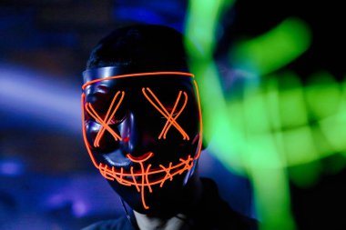 Kötü aydınlatılmış bir odada parlak renkli bir neon maske takan bir adam görülüyor..