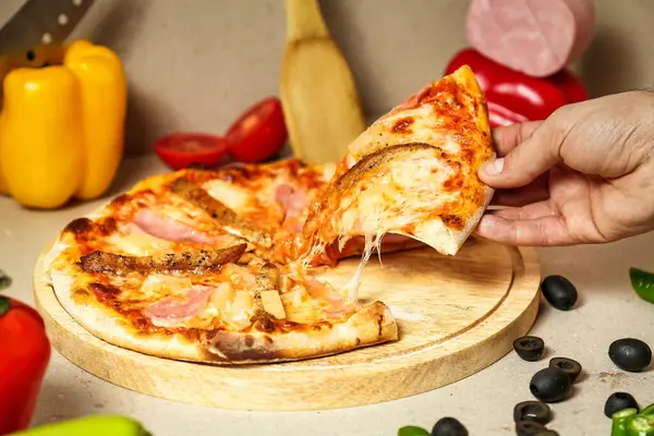 A persons hand reaches out to take a slice of pizza from a wooden cutting board.