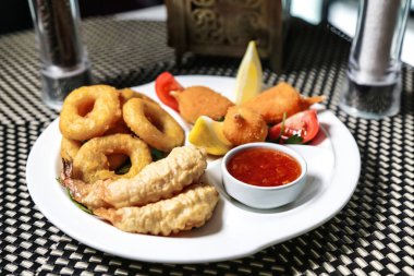 Tavuk kanatları, soğan halkaları ve patates kızartması gibi çeşitli kızarmış yiyeceklerle dolu beyaz bir tabak, yanında sosla servis edilir..