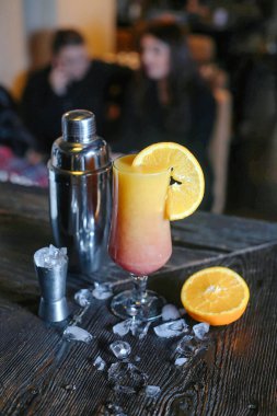 İçi portakal suyu dolu bir bardak masanın üstünde bir kokteyl kabının yanına yerleştirilmiş..