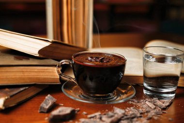 Bir fincan sıcak çikolata, bir bardak suyun yanındaki çay tabağında duruyor. Yanında da kitap ve çikolata parçacıkları var..